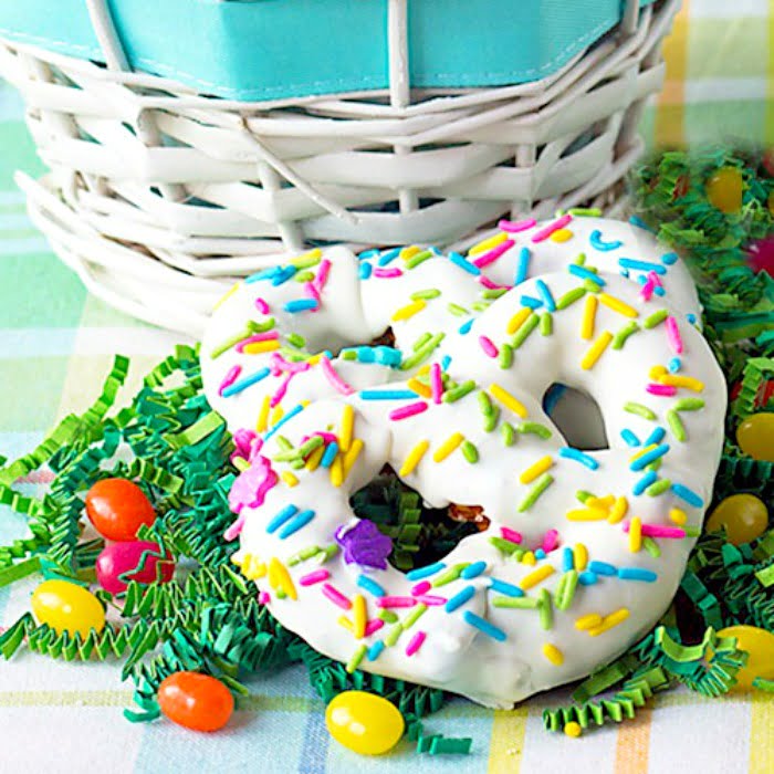 homemade white chocolate covered pretzels with Easter sprinkles and some jelly beans sitting on Easter grass in front of a white Easter basket